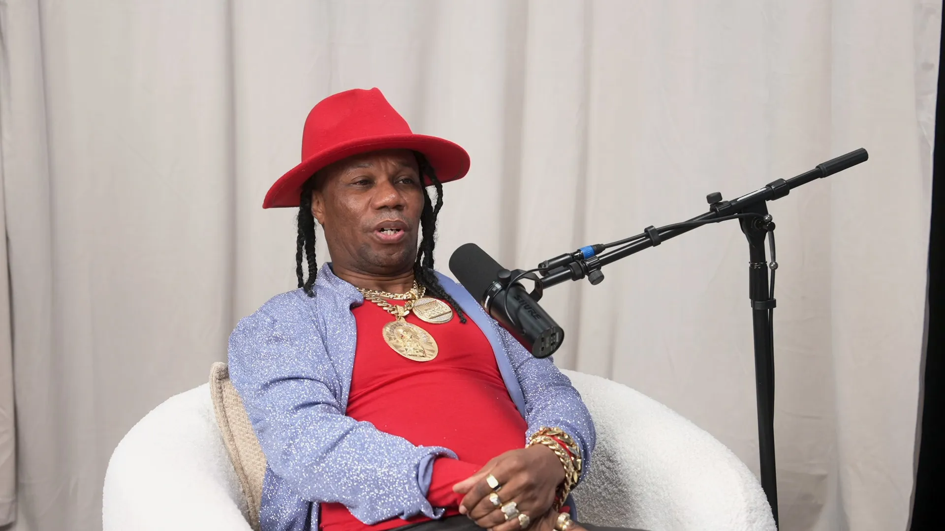 Chicho Severino, con sombrero rojo de ala ancha, camiseta roja, chaqueta azul brillante y cadenas doradas, habla ante un micrófono sentado en un sillón blanco