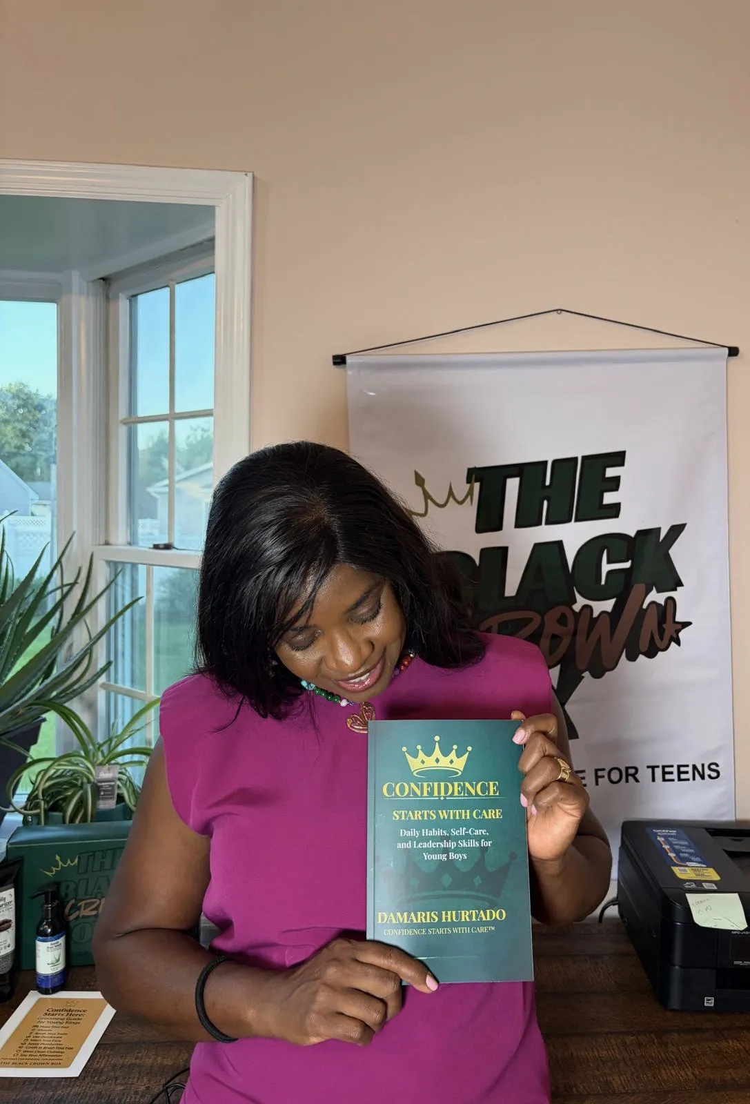 Damaris Hurtado muestra su libro Confidence Starts With Care junto a una ventana y un cartel de The Black Crown Box