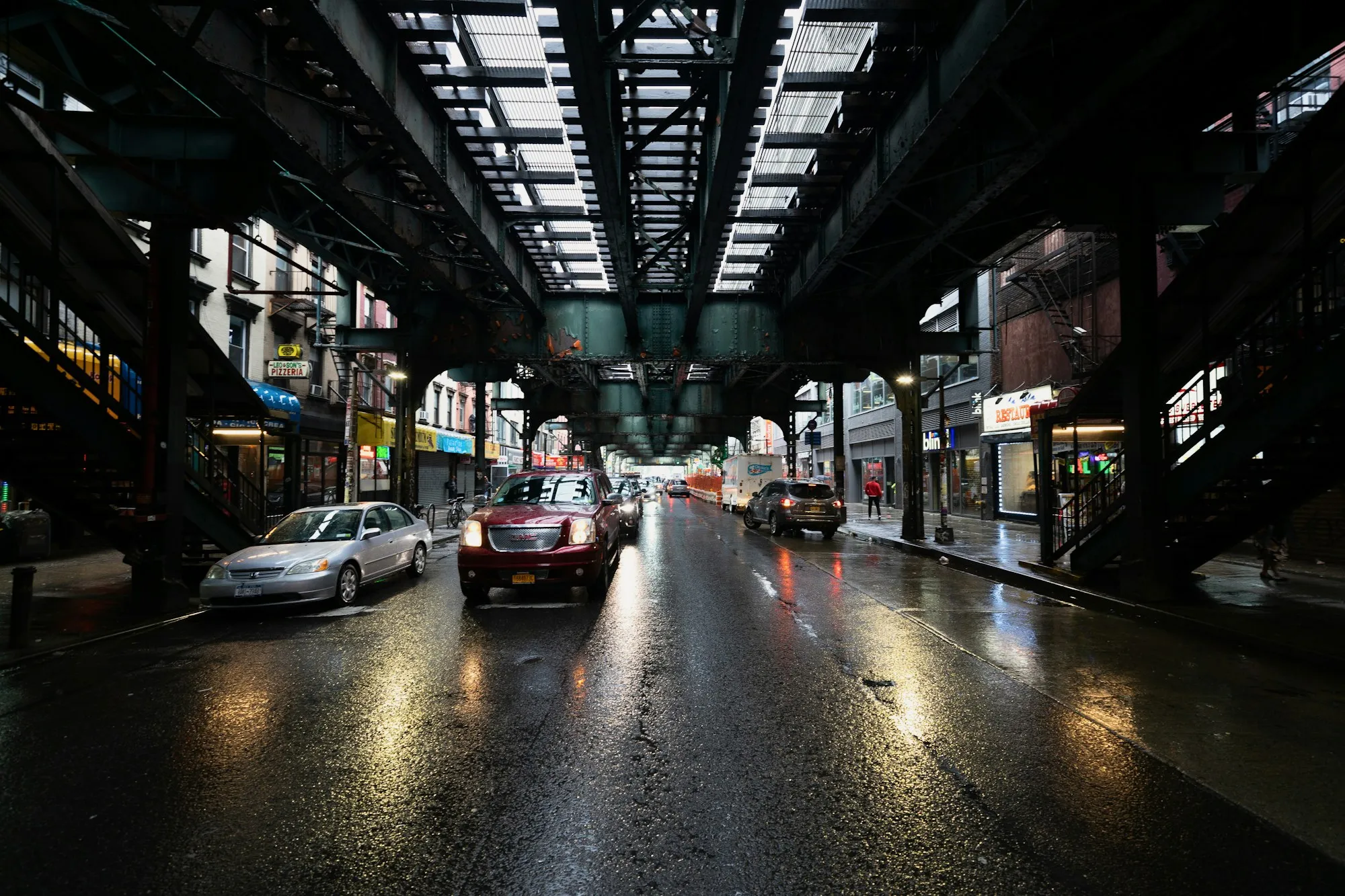 Calle mojada bajo las vías del tren elevado en Brooklyn, con coches y comercios a los lados